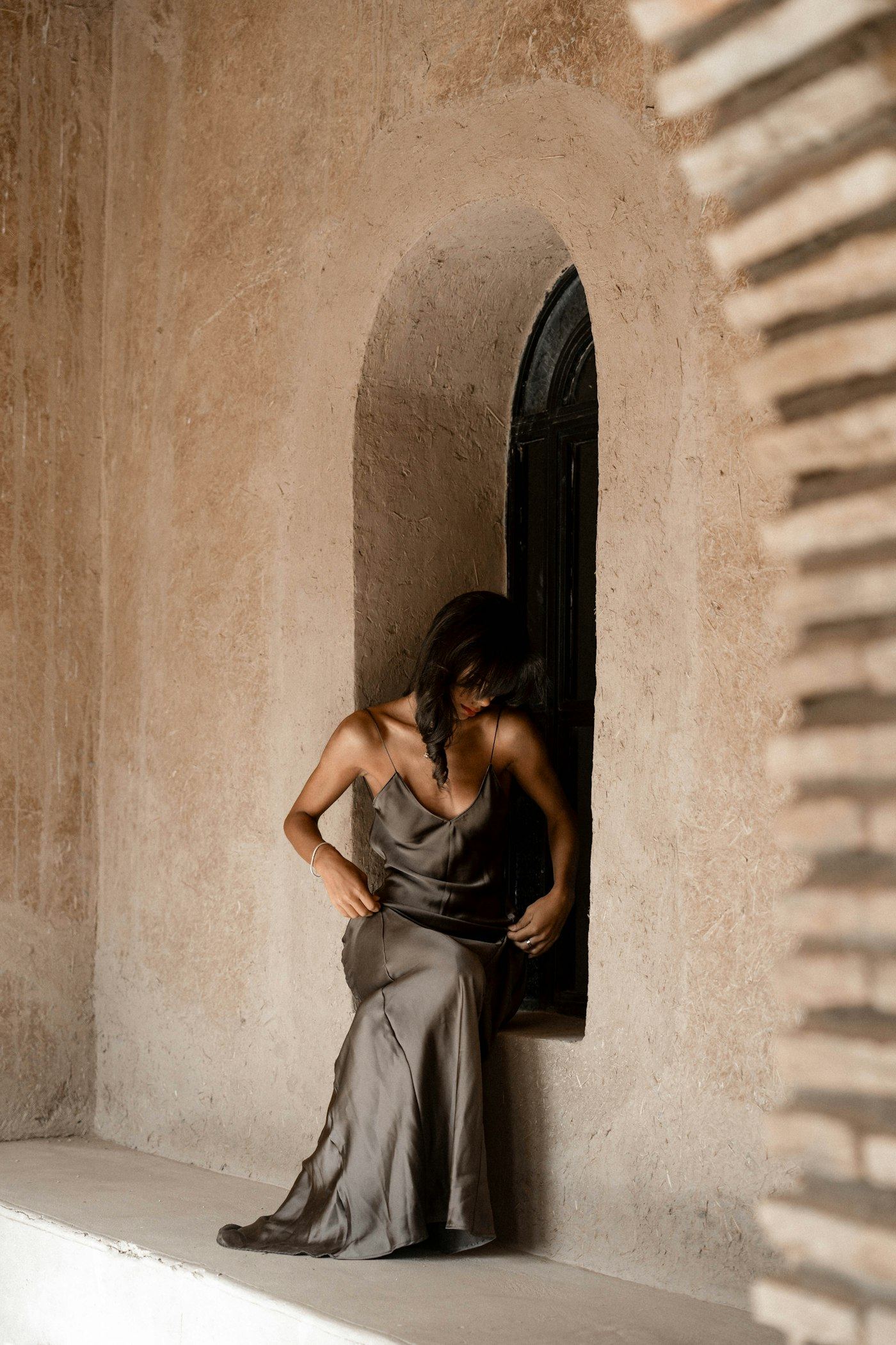 Woman in an olive-green dress with thin straps, seated in a deep arched window recess in a clay wall